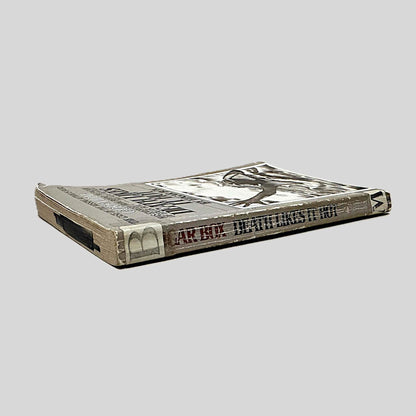 Close-up spine view of Back cover view of Death Likes It Hot by Edgar Box centered on a neutral light gray background. Spine shows creasing and chipping. There is a white sticker with a black capital "B" attached to the top end of the spine. Pages lay flat.