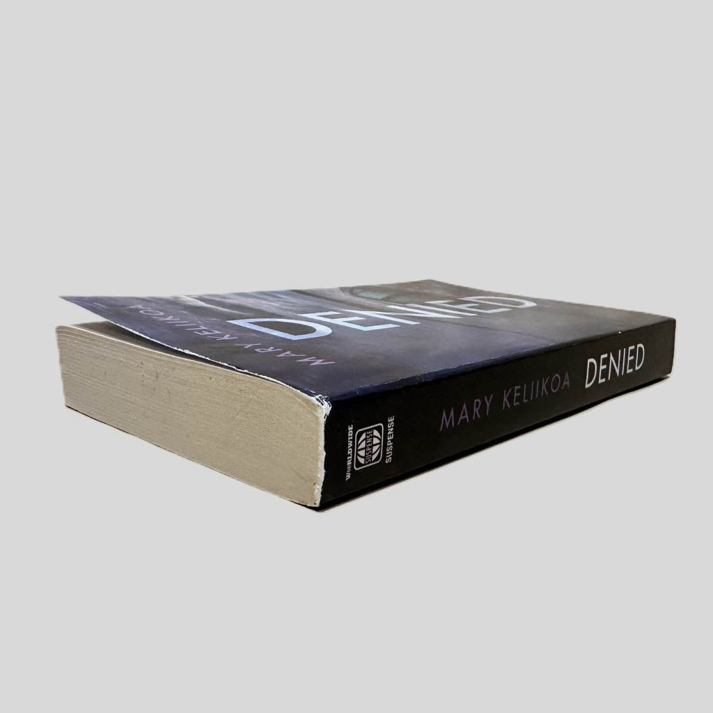Close-up spine view of Denied by Mary Keliikoa centered on a neutral light gray background. Spine shows minimal creasing. Pages lay flat. Some chipping at the top of the spine.