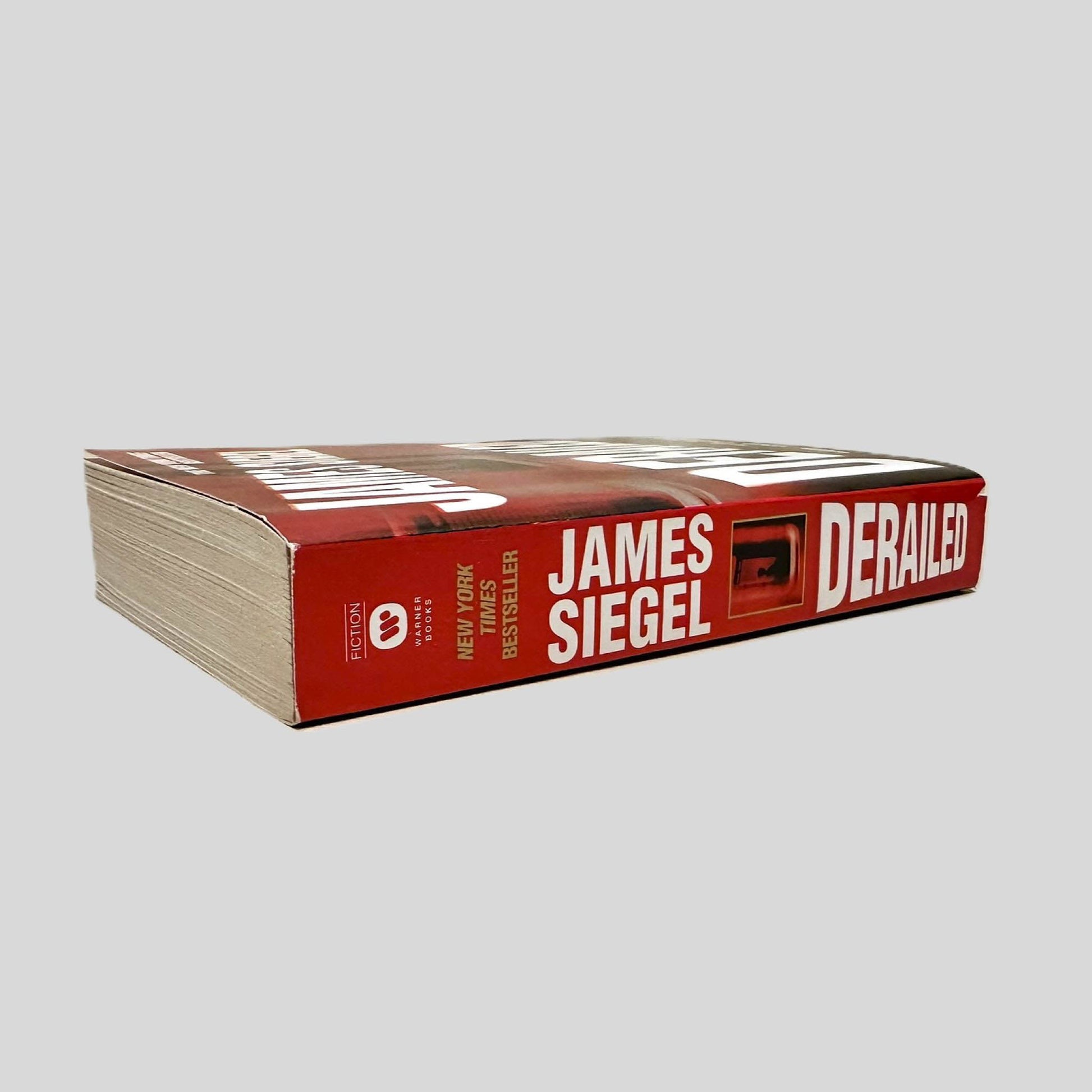 Close-up spine view of Derailed by James Siegel centered on a neutral light gray background. Spine is square and shows no creasing. Pages lay flat.