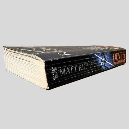 Close-up spine view of Devil's Plaything by Matt Richtel centered on a neutral light gray background. Spine shows creasing. Pages lay flat with the exception of dog-eared pages.