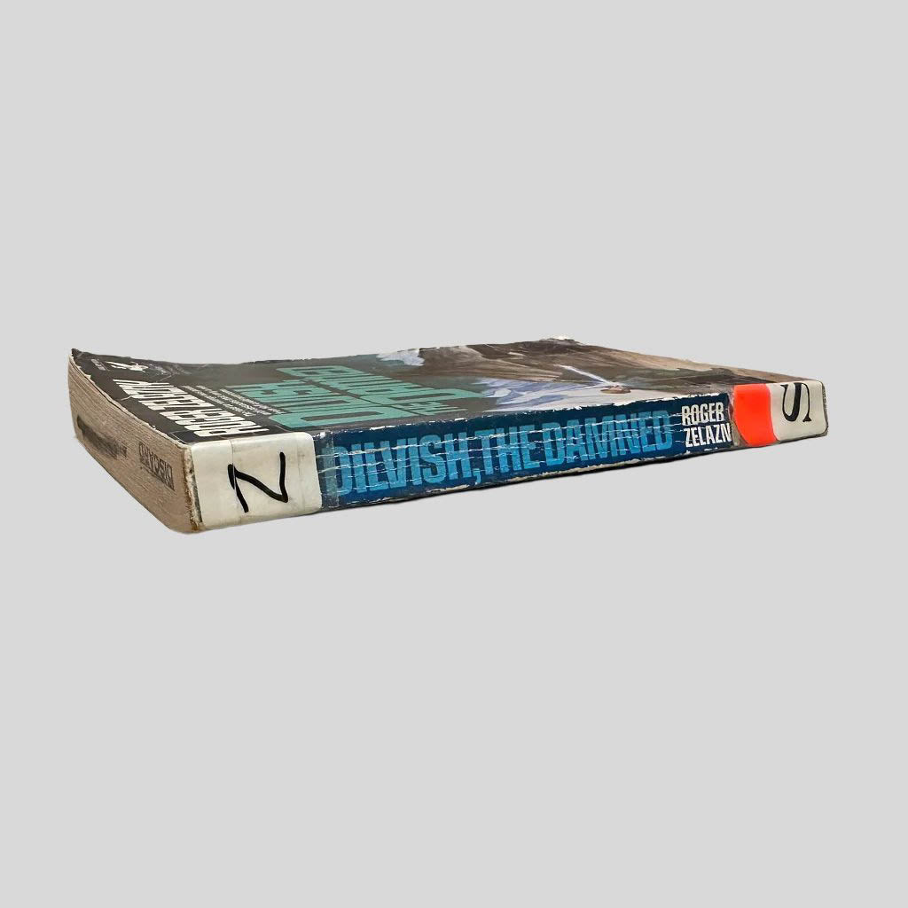 Close-up spine view of Dilvish, the Damned by Roger Zelazny centered on a neutral light gray background. Spine shows creasing. Library shelving stickers are attached at the top and lower end of the spine. Discard stamping marks on the top page edge. Pages lay flat.