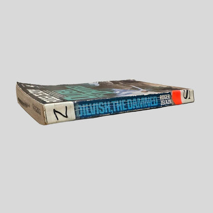 Close-up spine view of Dilvish, the Damned by Roger Zelazny centered on a neutral light gray background. Spine shows creasing. Library shelving stickers are attached at the top and lower end of the spine. Discard stamping marks on the top page edge. Pages lay flat.