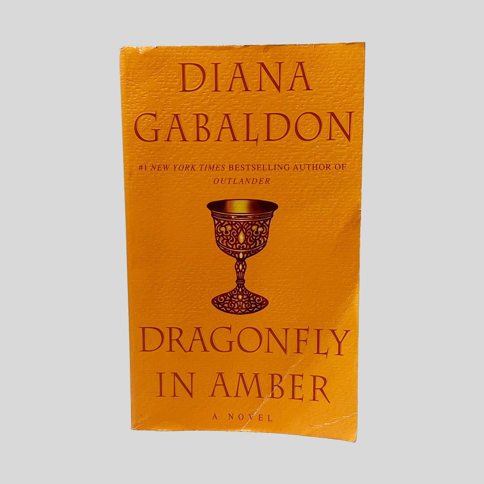 Front cover view of Dragonfly In Amber by Diana Gabaldon centered on a neutral light gray background. Chipping along the edges of the front cover from normal wear. Creasing along the bottom corner.