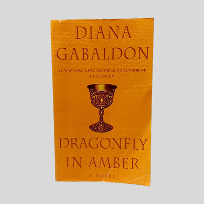 Front cover view of Dragonfly In Amber by Diana Gabaldon centered on a neutral light gray background. Chipping along the edges of the front cover from normal wear. Creasing along the bottom corner.