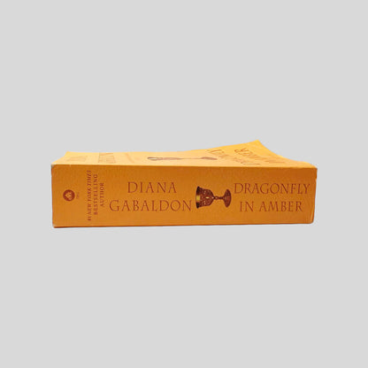 Close-up spine view of Outlander by Diana Gabaldon centered on a neutral light gray background. Spine shows minor bumping at the lower edge and no creasing. Spine is square and pages lay flat.
