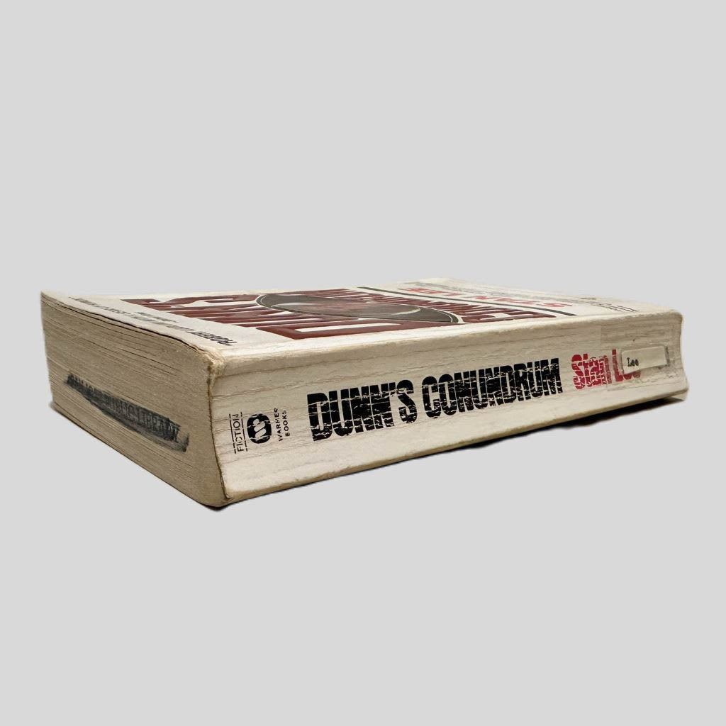 Close-up spine view of Dunn's Conundrum by Stan Lee centered on a neutral light gray background. Spine shows creasing. and chipping. Colored out black stamp on the top page edges. Library shelf sticker taped to lower spine. Pages lay flat.