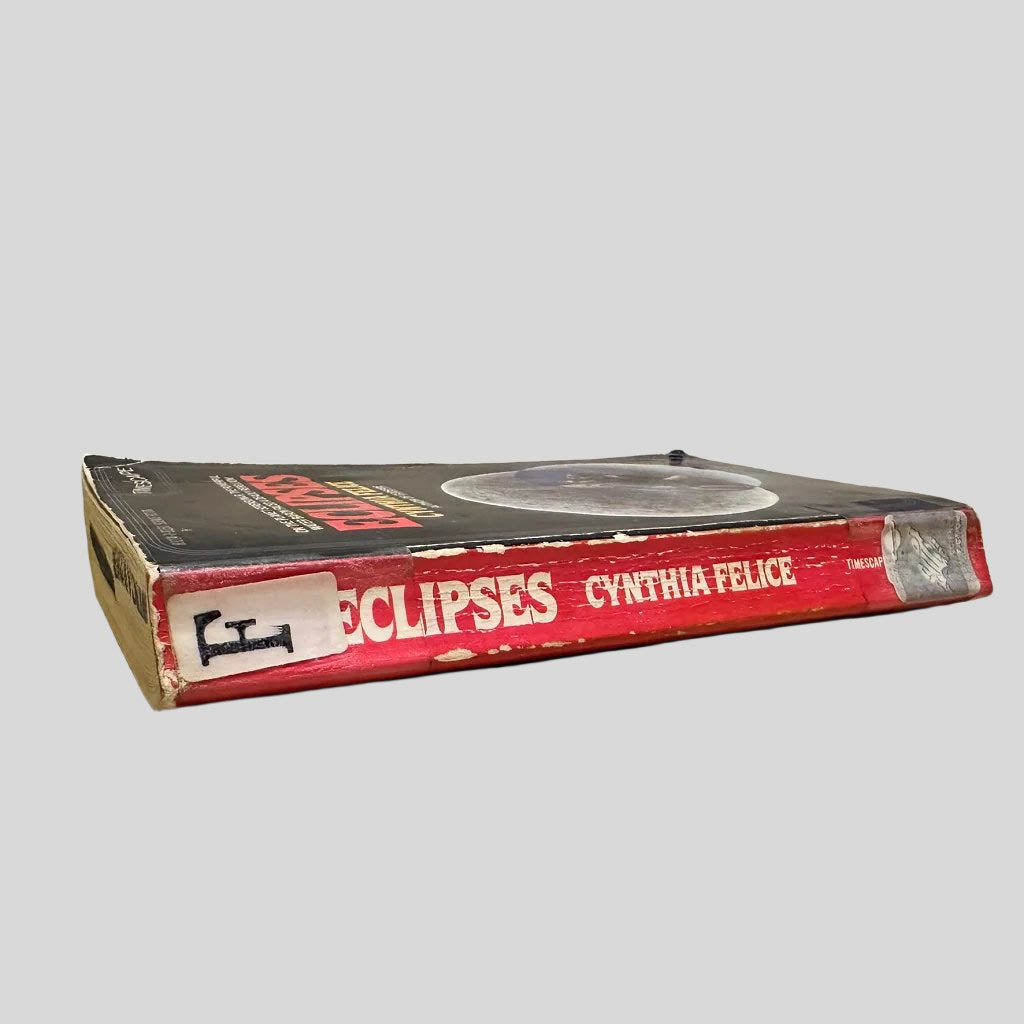 Close-up spine view of Eclipses by Cynthia Felice centered on a neutral light gray background. Spine shows creasing, chipping and cocking. Library shelving stickers taped to the top and lower spine. Pages lay flat. Discard stamp in black ink on the top page edges.