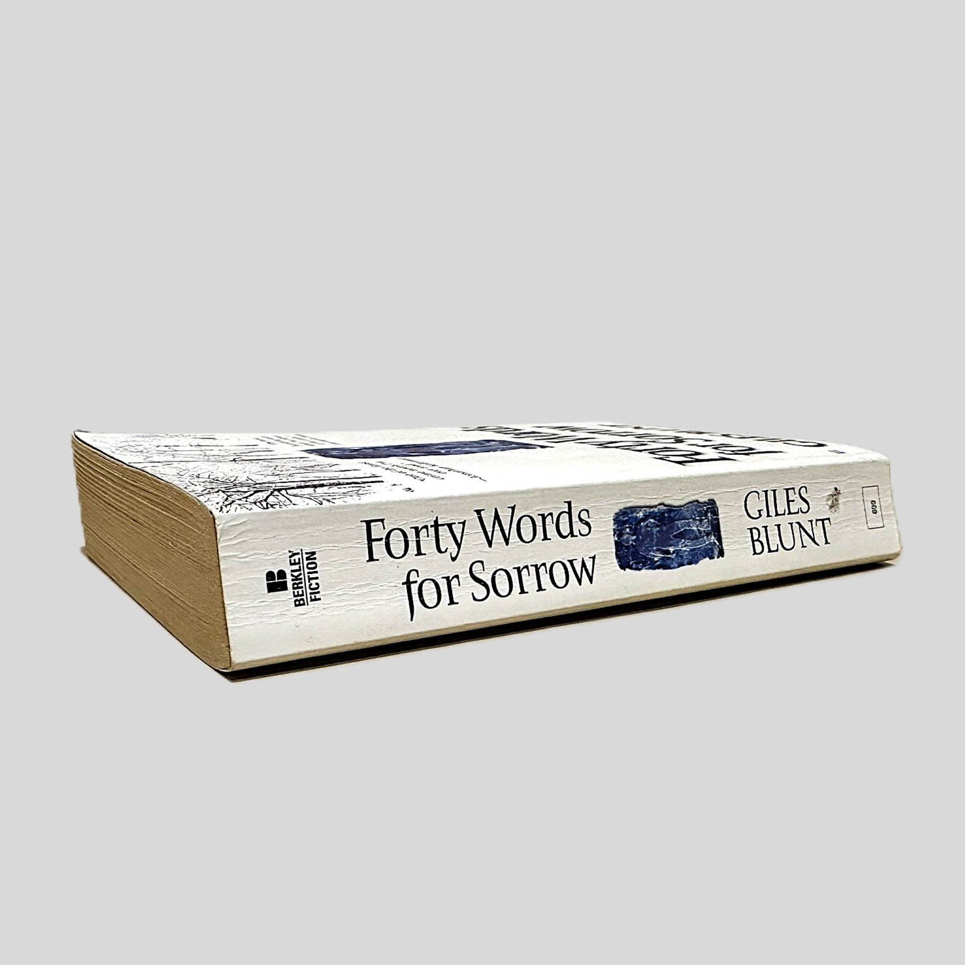 Close-up spine view of Forty Words for Sorrow by Giles Blunt centered on a neutral light gray background. Spine shows creasing. Pages lay flat.