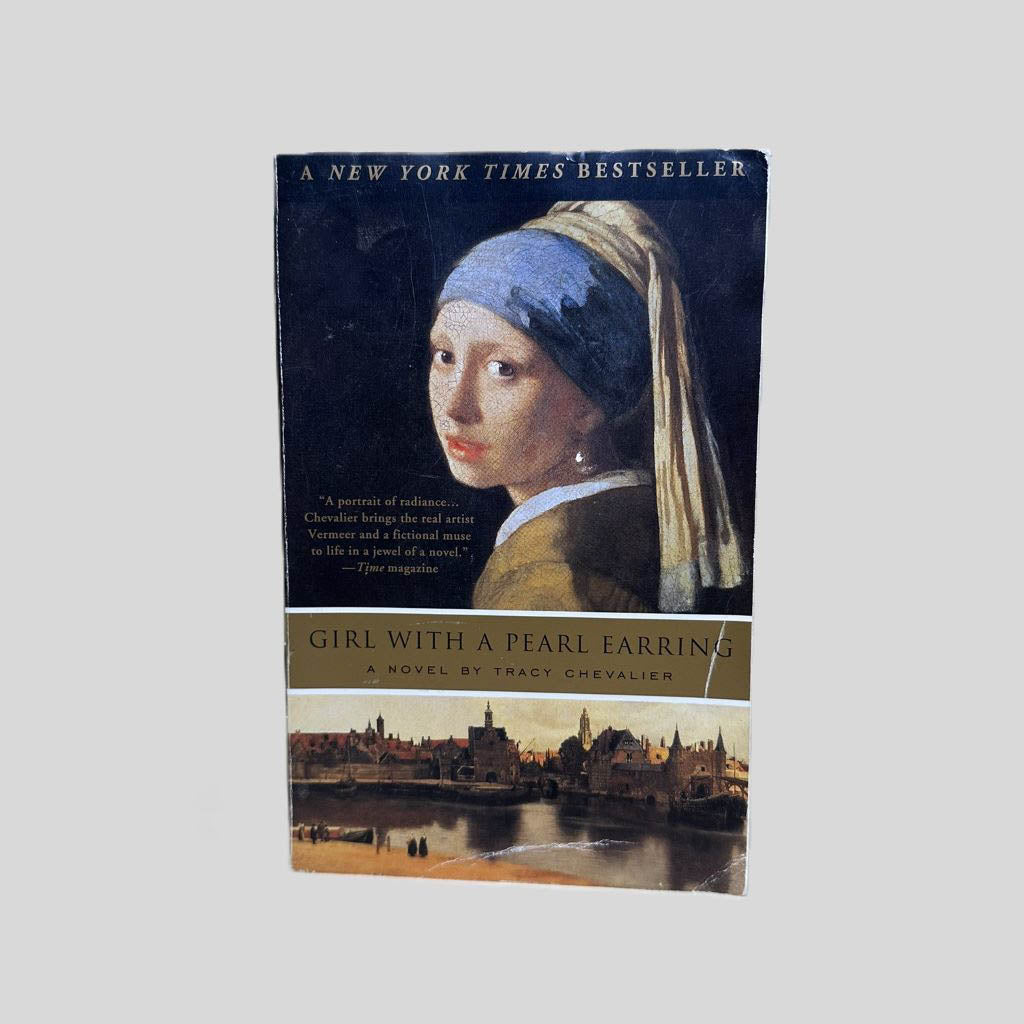 Front cover view of Girl with a Pearl Earring by Tracy Chevalier centered on a neutral light gray background. Creasing and chipping are present to the front cover. Minor water damage to some of the pages, leaving watermarking and some waves to the affected pages. Book is intact, text is not damaged. Cover art is vibrant.