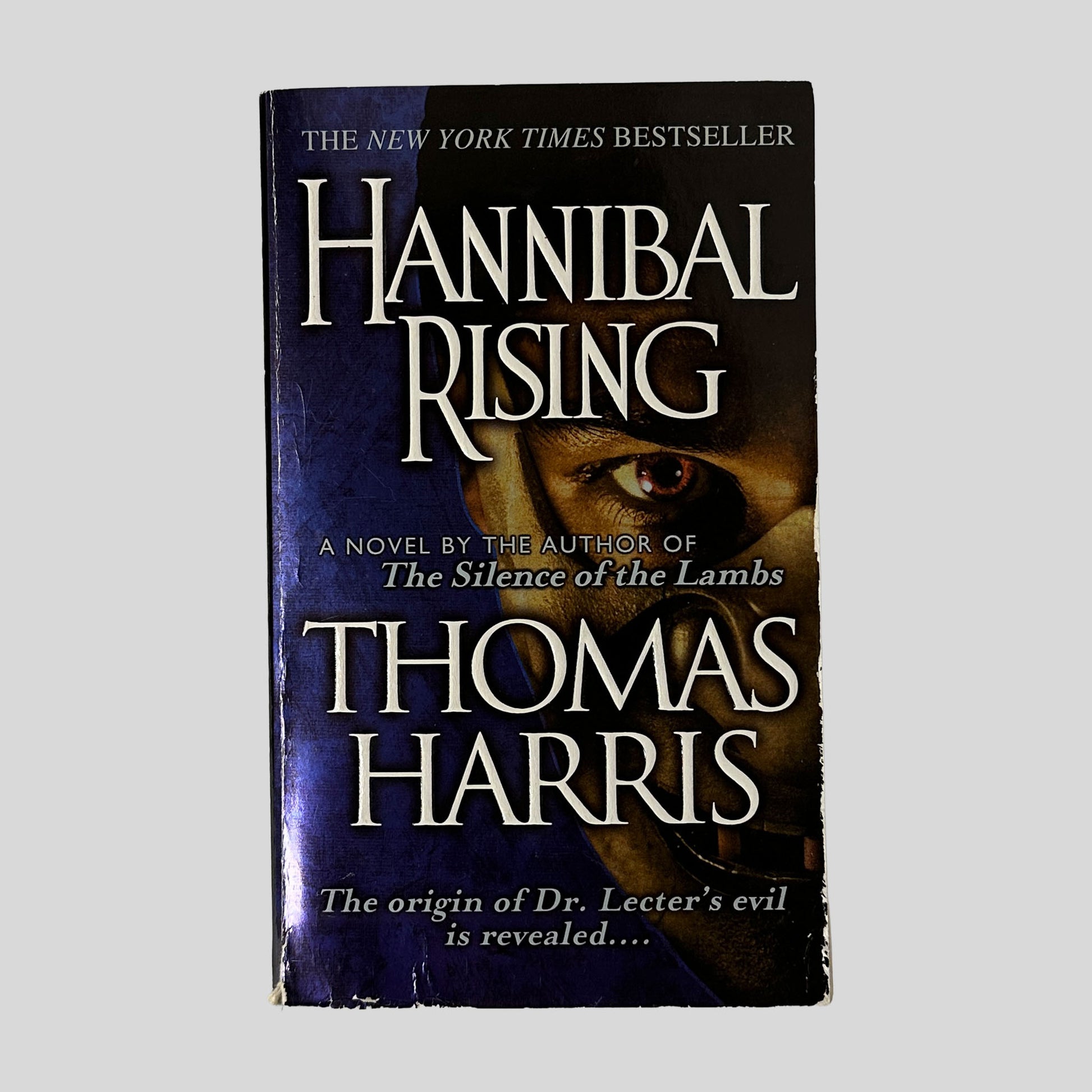 The front cover of the paperback Hannibal Rising by Thomas Harris, featuring a dark blue and black design with a close-up of a single, intense human eye peering through a mask.