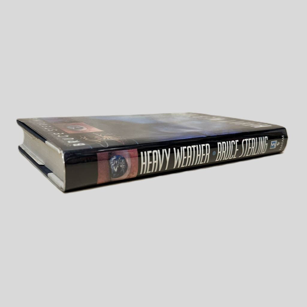 Close-up spine view of Heavy Weather by Bruce Sterling centered on a neutral light gray background. Dust jacket is present and undamaged. Spine shows no creasing. Pages lay flat.