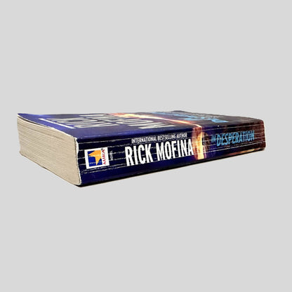 Close-up spine view of In Desperation by Rick Mofina centered on a neutral light gray background. Spine shows creasing and chipping at the top of the spine.