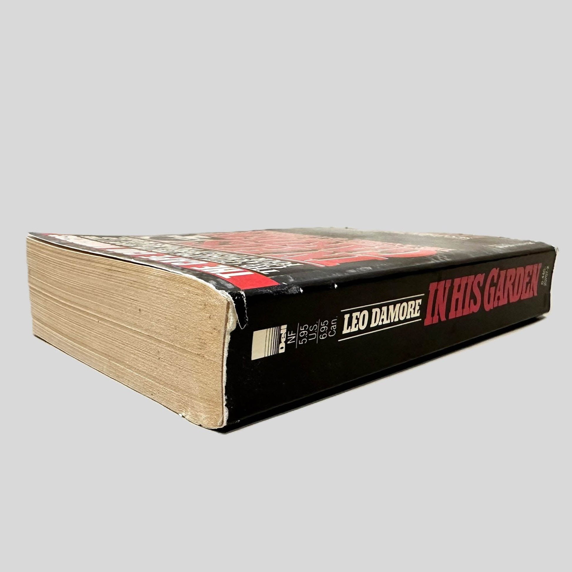 Close-up spine view of In His Garden by Leo Damore centered on a neutral light gray background. Spine shows minimal creasing and bumping on along the top spine edge.