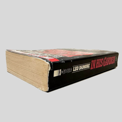 Close-up spine view of In His Garden by Leo Damore centered on a neutral light gray background. Spine shows minimal creasing and bumping on along the top spine edge.