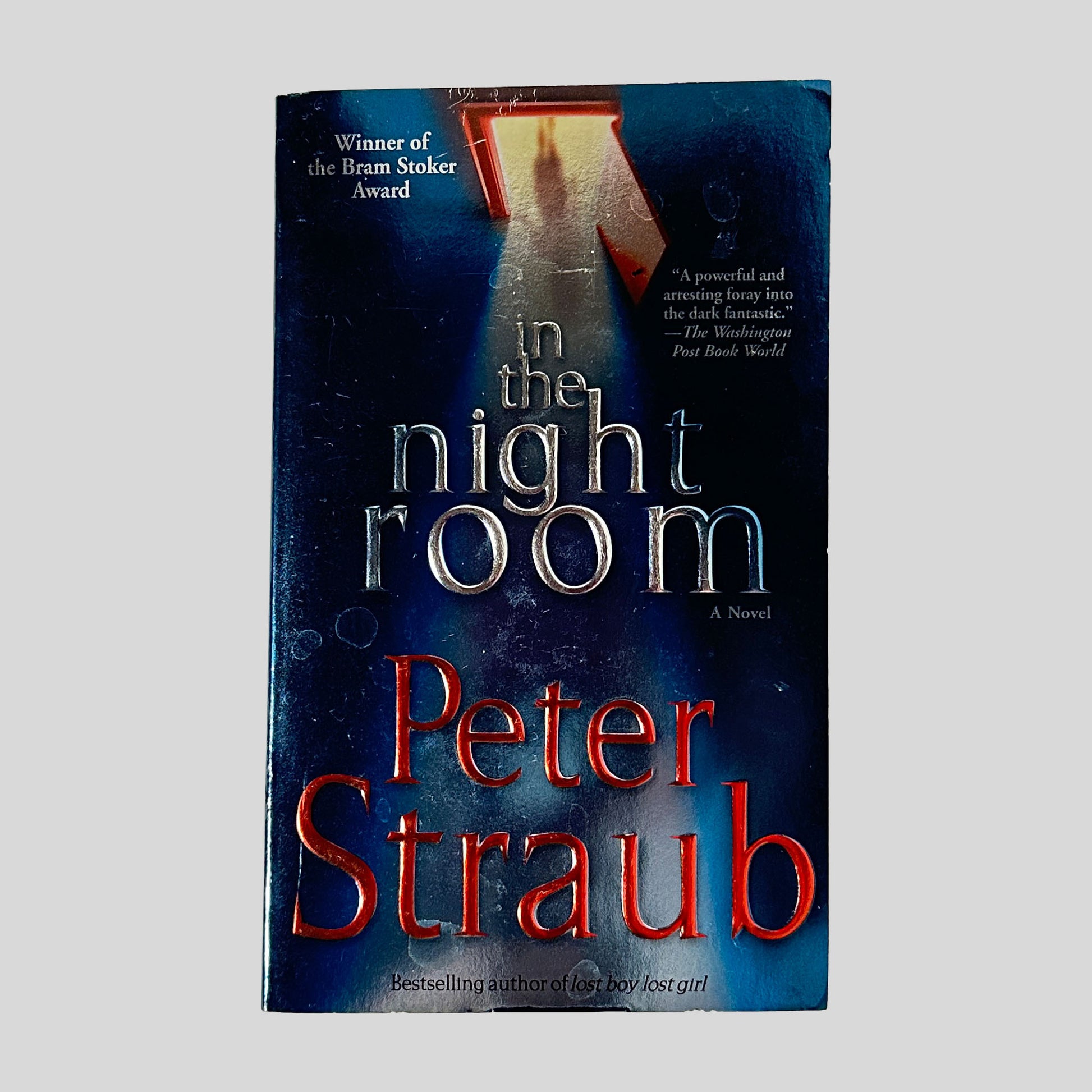 The dark blue front cover of the paperback In the Night Room by Peter Straub, featuring silver and red embossed title text below an image of a shadowy figure in a doorway.