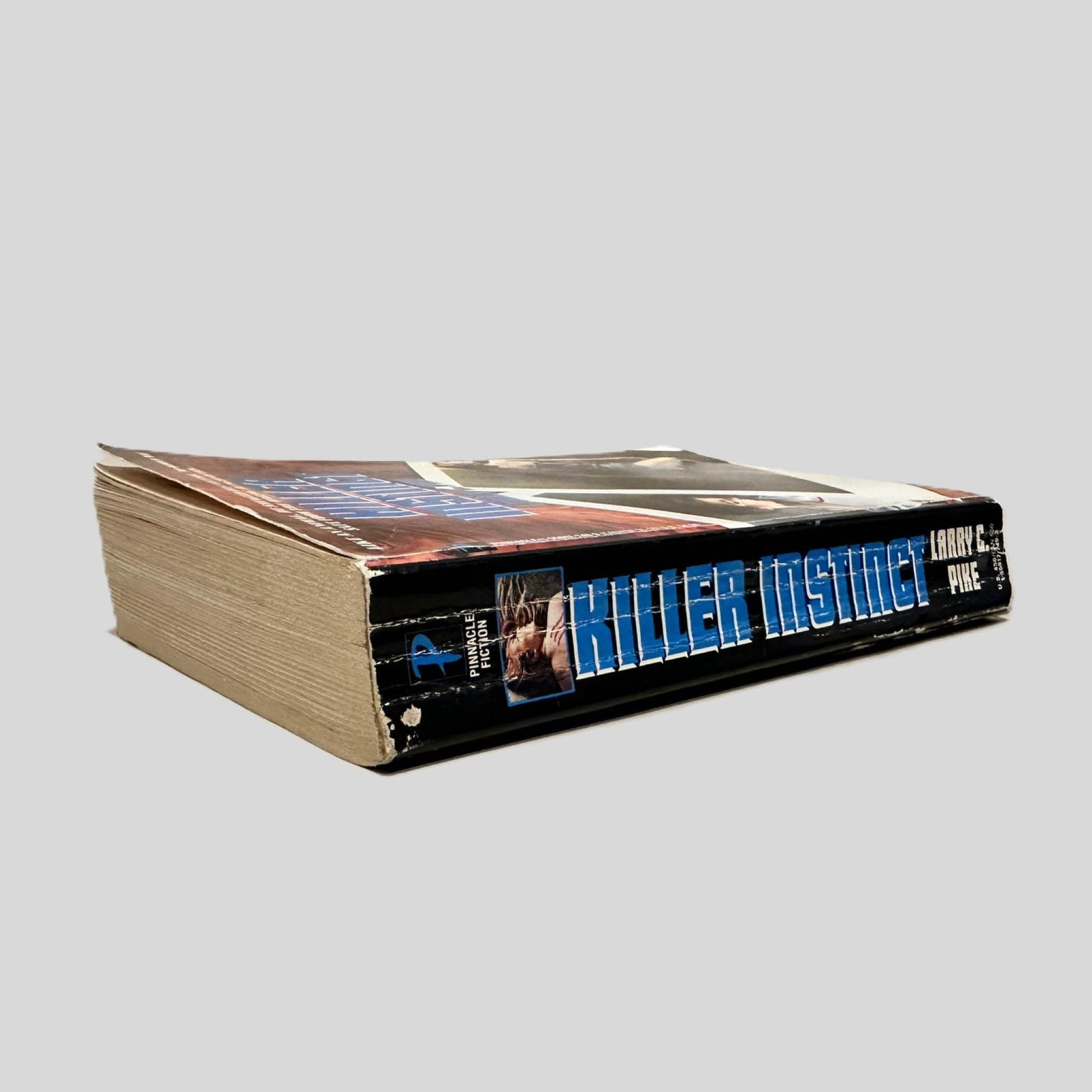 Close-up spine view of Killer Instinct by Larry C Pike centered on a neutral light gray background. Spine shows creasing. Pages lay flat.