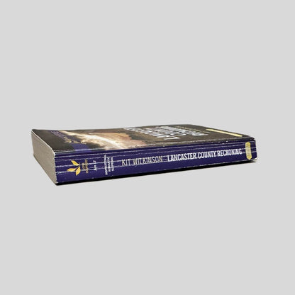 Close-up spine view of Lancaster County Reckoning by Kit Wilkinson centered on a neutral light gray background.  Large print edition. Spine shows creasing. Pages lay flat.