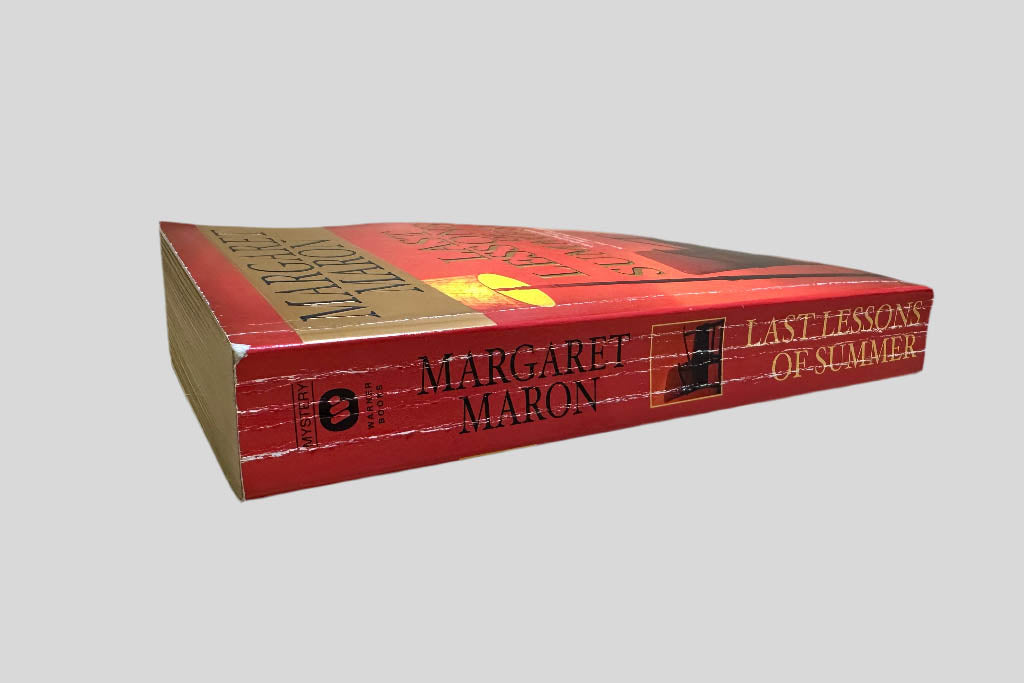 Close-up spine view of Last Lessons of Summer by Margaret Maron centered on a neutral light gray background. Spine shows creasing. Minor chip at the top corner of the spine. Pages lay flat.