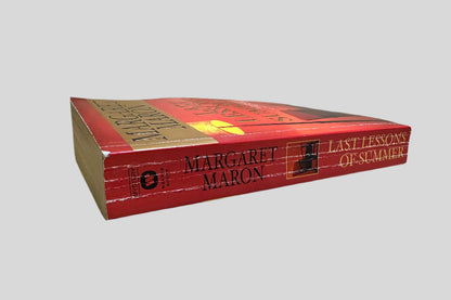 Close-up spine view of Last Lessons of Summer by Margaret Maron centered on a neutral light gray background. Spine shows creasing. Minor chip at the top corner of the spine. Pages lay flat.