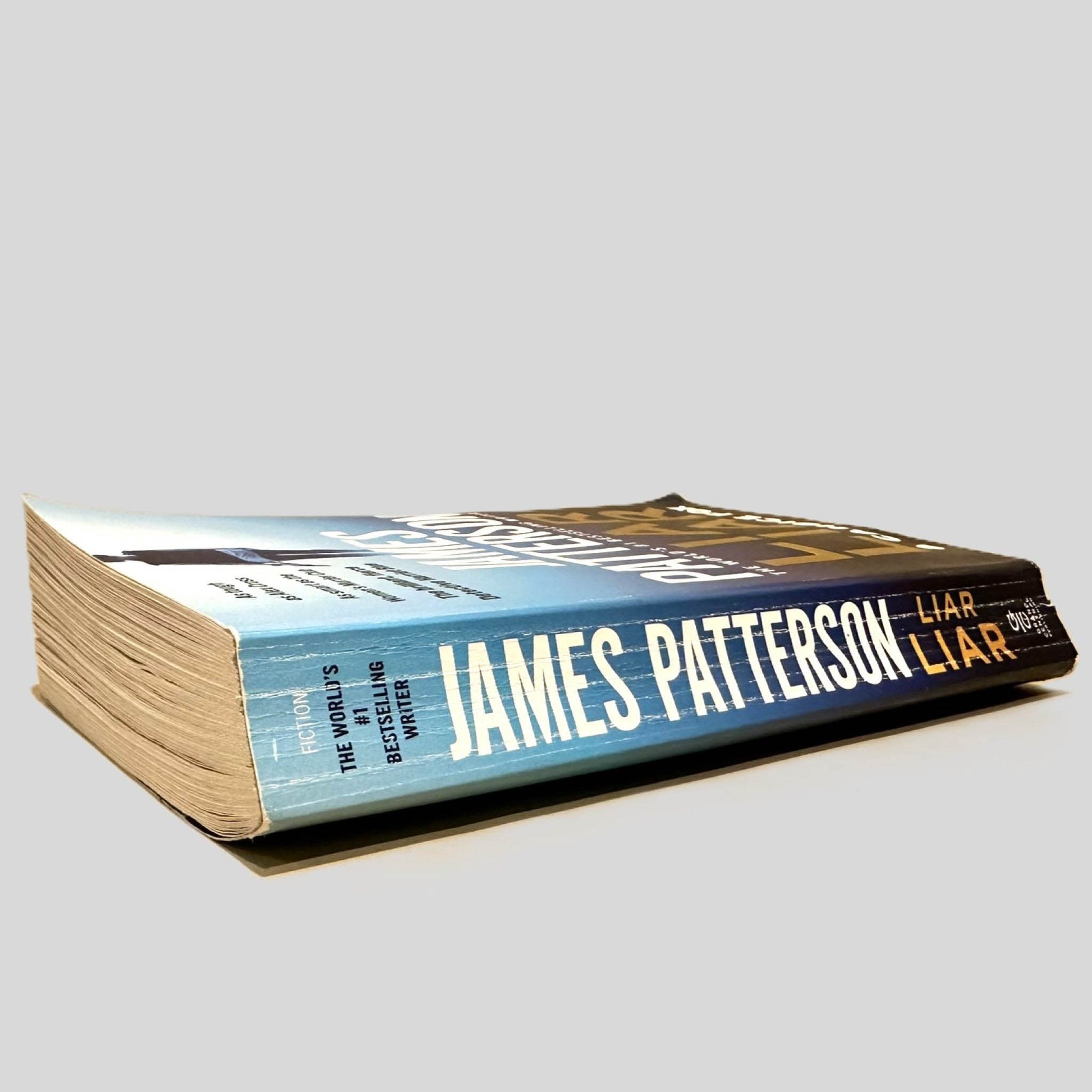 Close-up spine view of Liar Liar by James Patterson & Candice Fox centered on a neutral light gray background. Spine shows creasing and cocking.