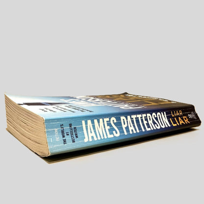 Close-up spine view of Liar Liar by James Patterson & Candice Fox centered on a neutral light gray background. Spine shows creasing and cocking.
