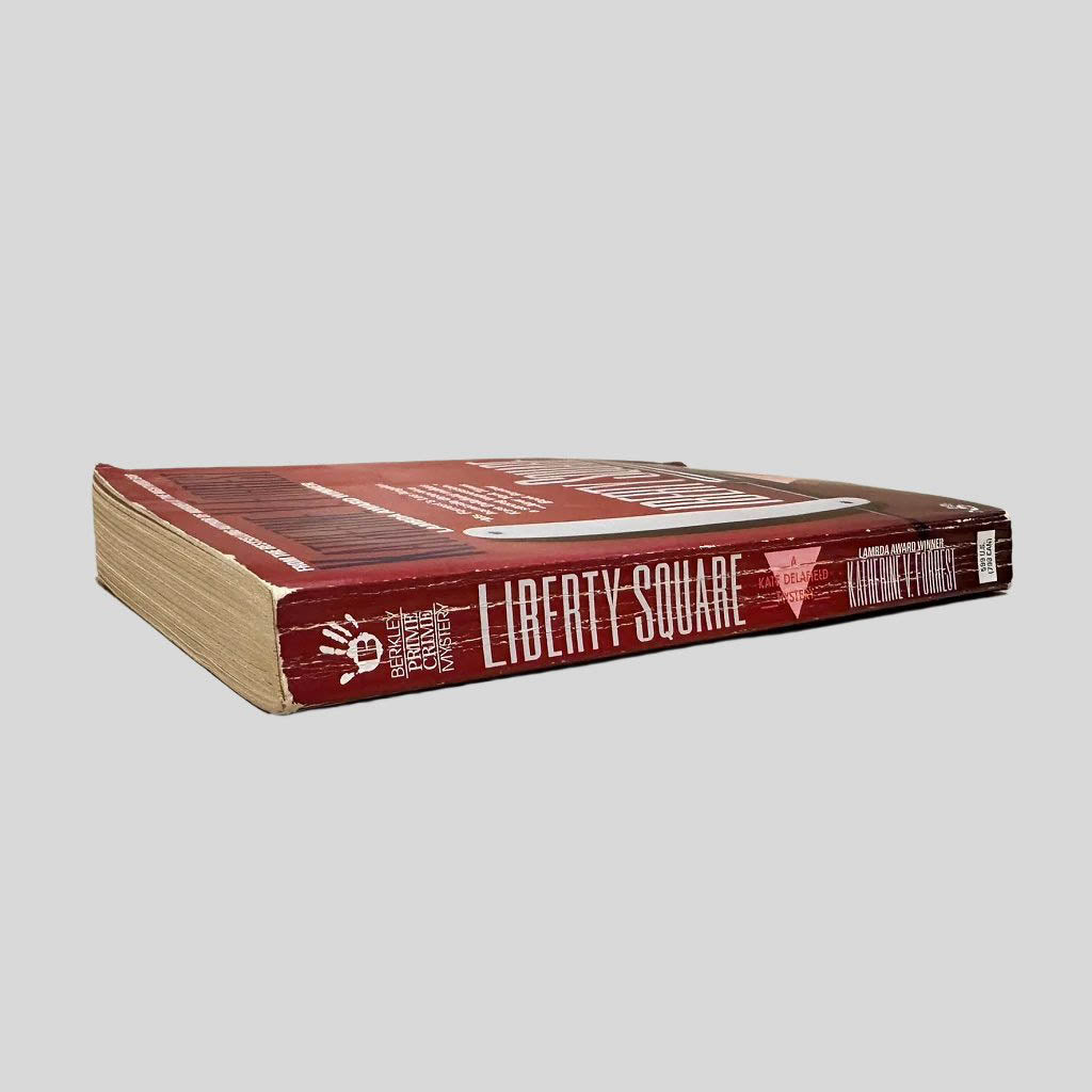Close-up spine view of Liberty Square by Katherine V Forrest centered on a neutral light gray background. Spine shows creasing. Pages lay flat.