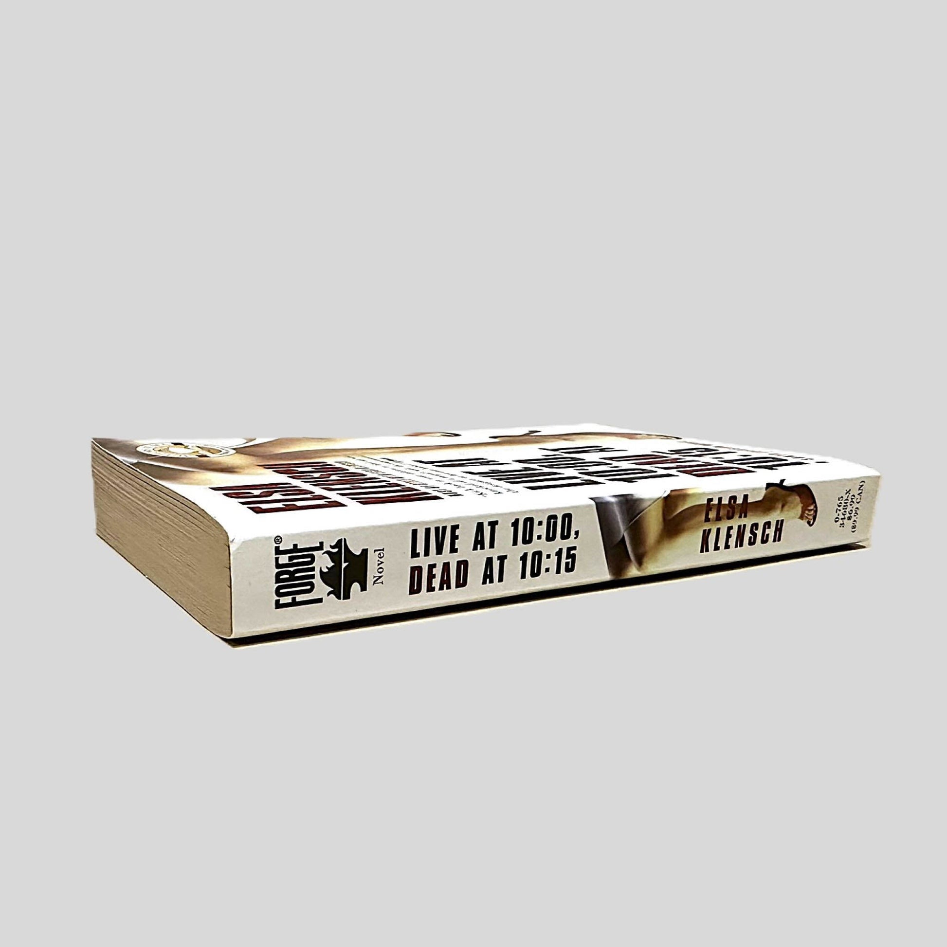 Close-up spine view of Live at 10:00, Dead at 10:15 by Elsa Klensch centered on a neutral light gray background. Spine is square with no creasing. Pages lay flat.