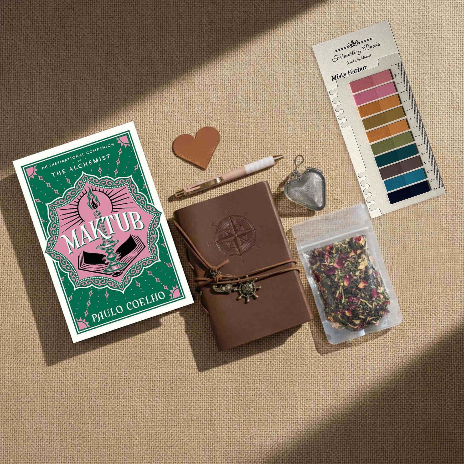 Overhead view of the "Maktub" Ultimate Literary Collection featuring the hardcover book by Paulo Coelho, a brown leather-bound journal with compass embossing, a heart-shaped tea infuser, "Self Care" herbal tea, and a "Misty Harbor" annotation tab palette on a burlap surface.