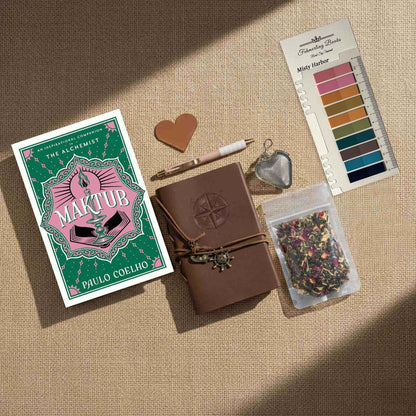 Overhead view of the "Maktub" Ultimate Literary Collection featuring the hardcover book by Paulo Coelho, a brown leather-bound journal with compass embossing, a heart-shaped tea infuser, "Self Care" herbal tea, and a "Misty Harbor" annotation tab palette on a burlap surface.