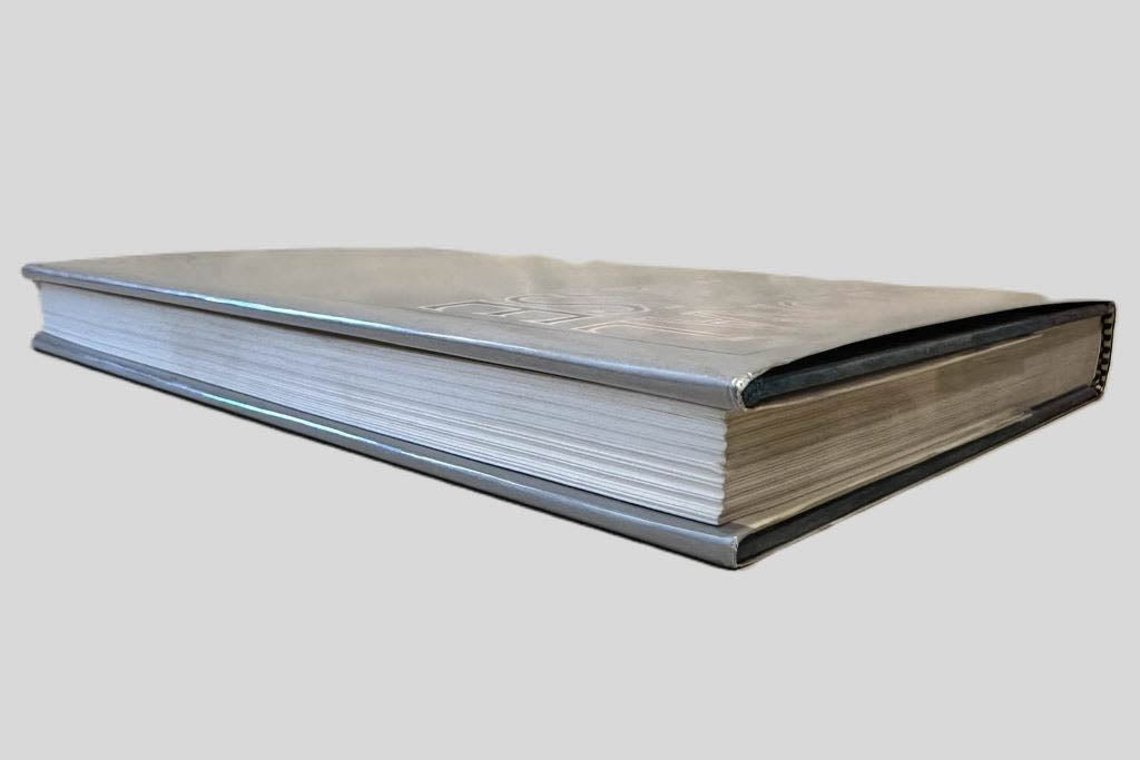 Close-up page edge view of National Defense by James Fallows centered on a neutral light gray background. Dust jacket is present and shows no damage. Spine shows no creasing. Pages lay flat.