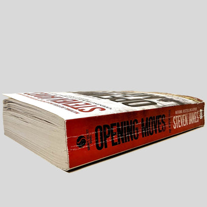 Close-up spine view of Opening Moves by Steven James centered on a neutral light gray background. Spine shows creasing. Page corners at the top of the book show bumping. but otherwise lay flat.