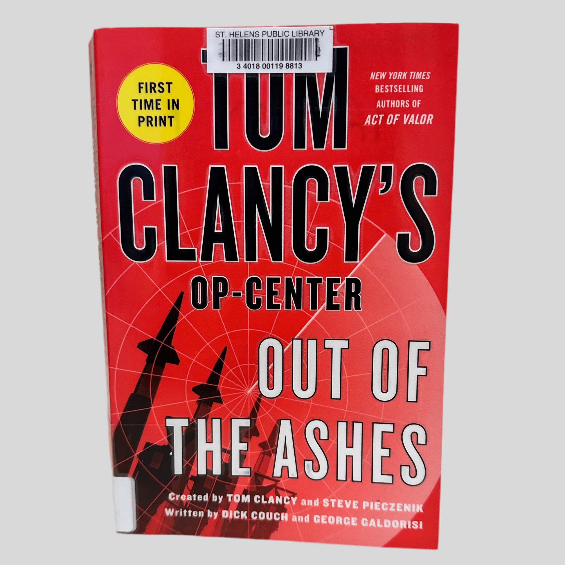 Front cover view of Out of the Ashes by Tom Clancy centered on a neutral light gray background. St. Helens Public Library barcode sticker is present, centered at the top of the book, covering part of the author's name. 