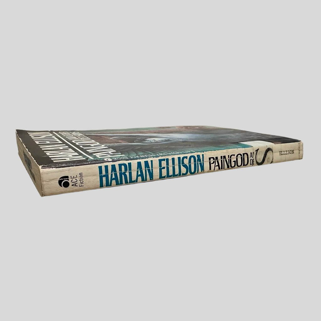 Close-up spine view of Paingod and Other Delusions by Harlan Ellison centered on a neutral light gray background. Spine is square and shows minimal creasing. Library shelving stickers attached to the lower end of the spine. Pages lay flat.