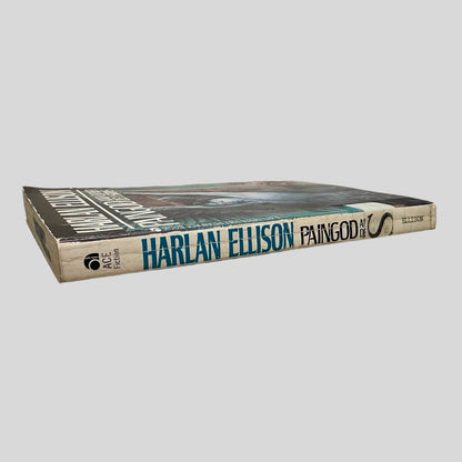 Close-up spine view of Paingod and Other Delusions by Harlan Ellison centered on a neutral light gray background. Spine is square and shows minimal creasing. Library shelving stickers attached to the lower end of the spine. Pages lay flat.
