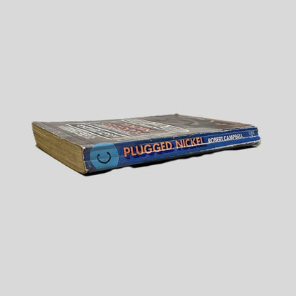 Close-up spine view of Plugged Nickel by Robert Campbell centered on a neutral light gray background. Spine shows creasing. Pages lay flat. There is a round blue sticker with the letter "C" in black ink on it with clear tape holding in place for library cataloging. 