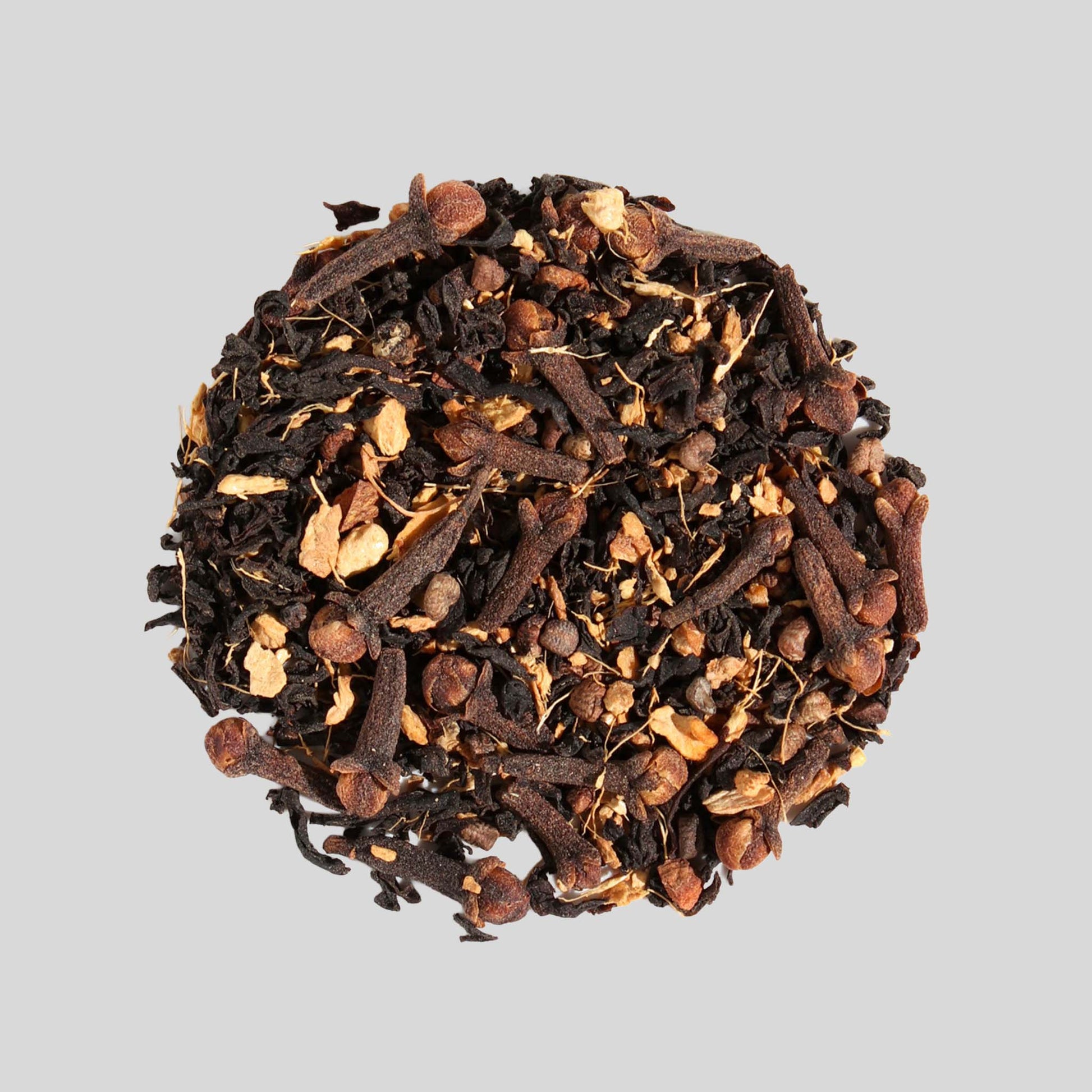 A close-up, top-down view of a small, circular mound of "Full Moon Chai" loose-leaf tea. The blend clearly shows dark tea leaves mixed with large, woody pieces of cinnamon bark and several whole cloves against a neutral grey background.