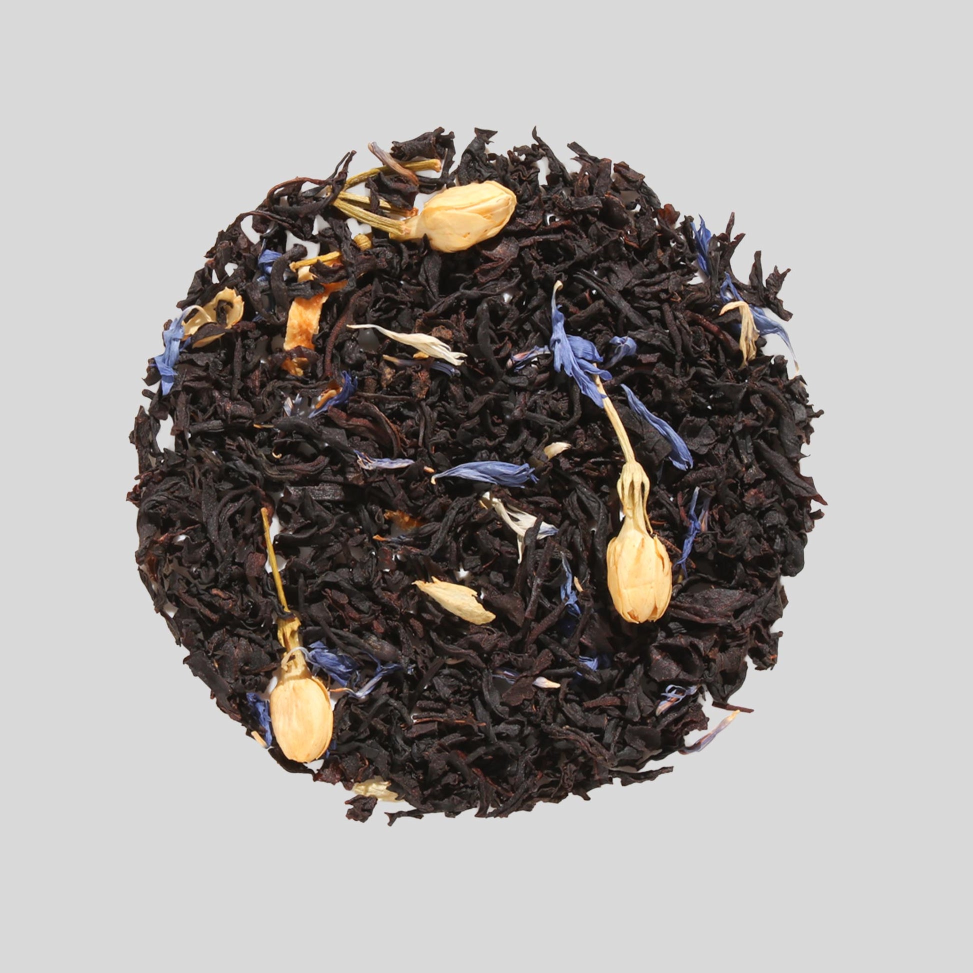 Plum Deluxe "Creme Brulée Grey" loose-leaf black tea featuring whole jasmine buds and blue cornflower petals arranged in a circle on a neutral background.