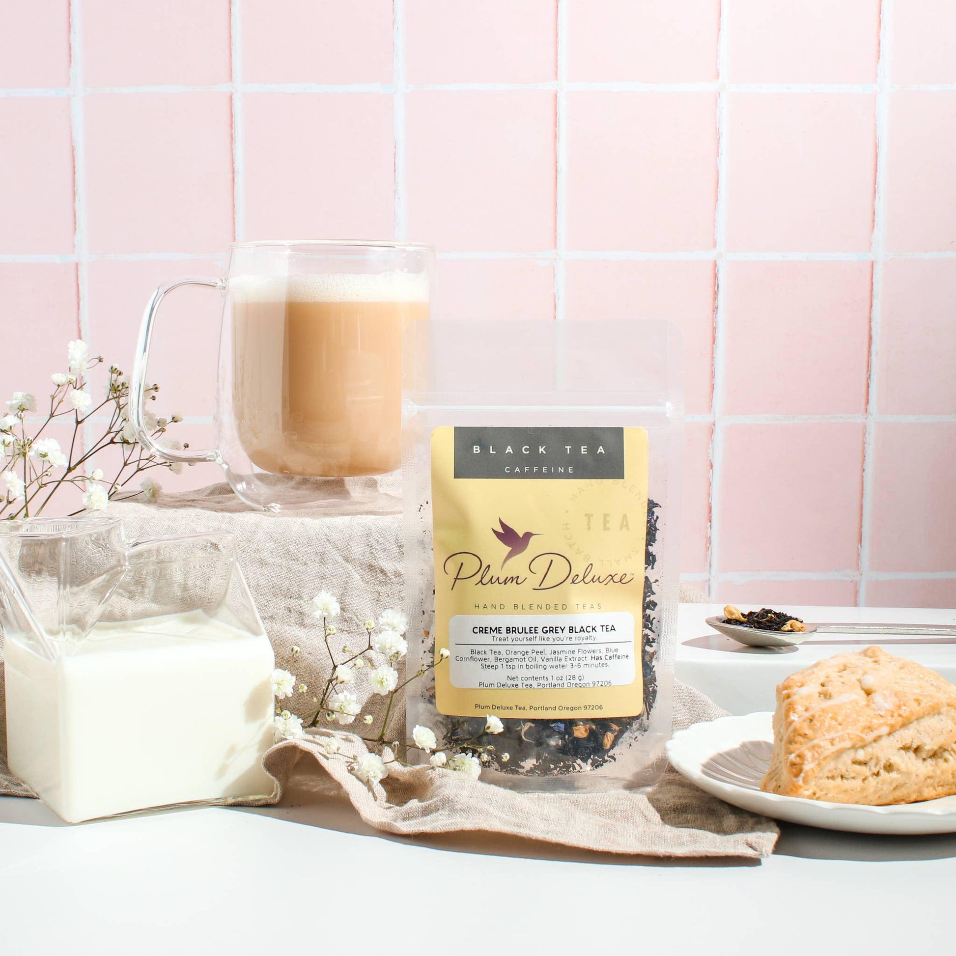 A 1oz package of Plum Deluxe "Creme Brulée Grey" black tea displayed with a frothed tea latte in a glass mug, a scone, and white baby's breath flowers against a light pink tiled backdrop.