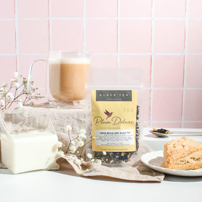 A 1oz package of Plum Deluxe "Creme Brulée Grey" black tea displayed with a frothed tea latte in a glass mug, a scone, and white baby's breath flowers against a light pink tiled backdrop.