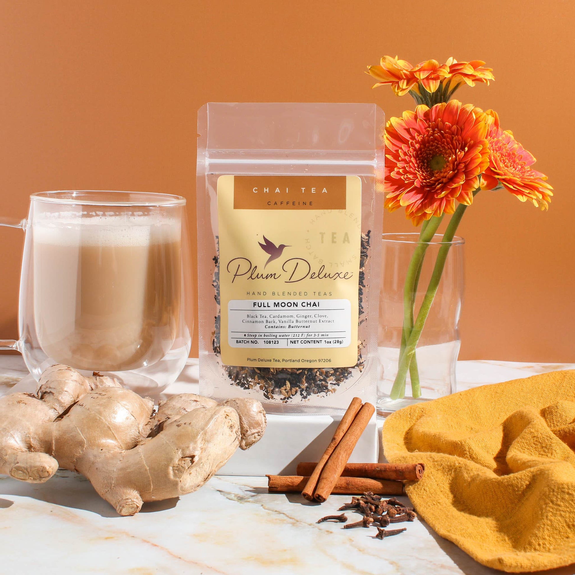 A clear 1oz package of Plum Deluxe "Full Moon Chai" tea stands on a marble surface. Surrounding the package is a glass mug filled with creamy, frothed tea, fresh ginger root, cinnamon sticks, and whole cloves. In the background, a small glass vase holds vibrant orange flowers against a warm, orange-toned backdrop.