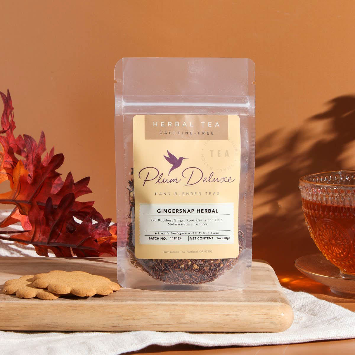 A 1oz package of Plum Deluxe "Gingersnap Herbal" tea featuring red rooibos and ginger, displayed on a wooden board with two gingersnap cookies, a glass of brewed tea, and red autumn leaves against a terracotta background.