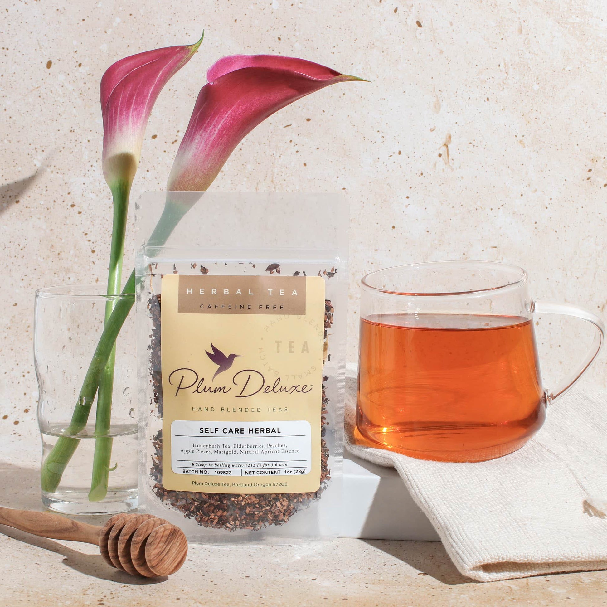A 1oz package of Plum Deluxe "Self Care" herbal tea featuring apricot and elderberry, displayed with a glass of brewed tea, pink calla lilies, and a wooden honey dipper on a neutral textured surface.