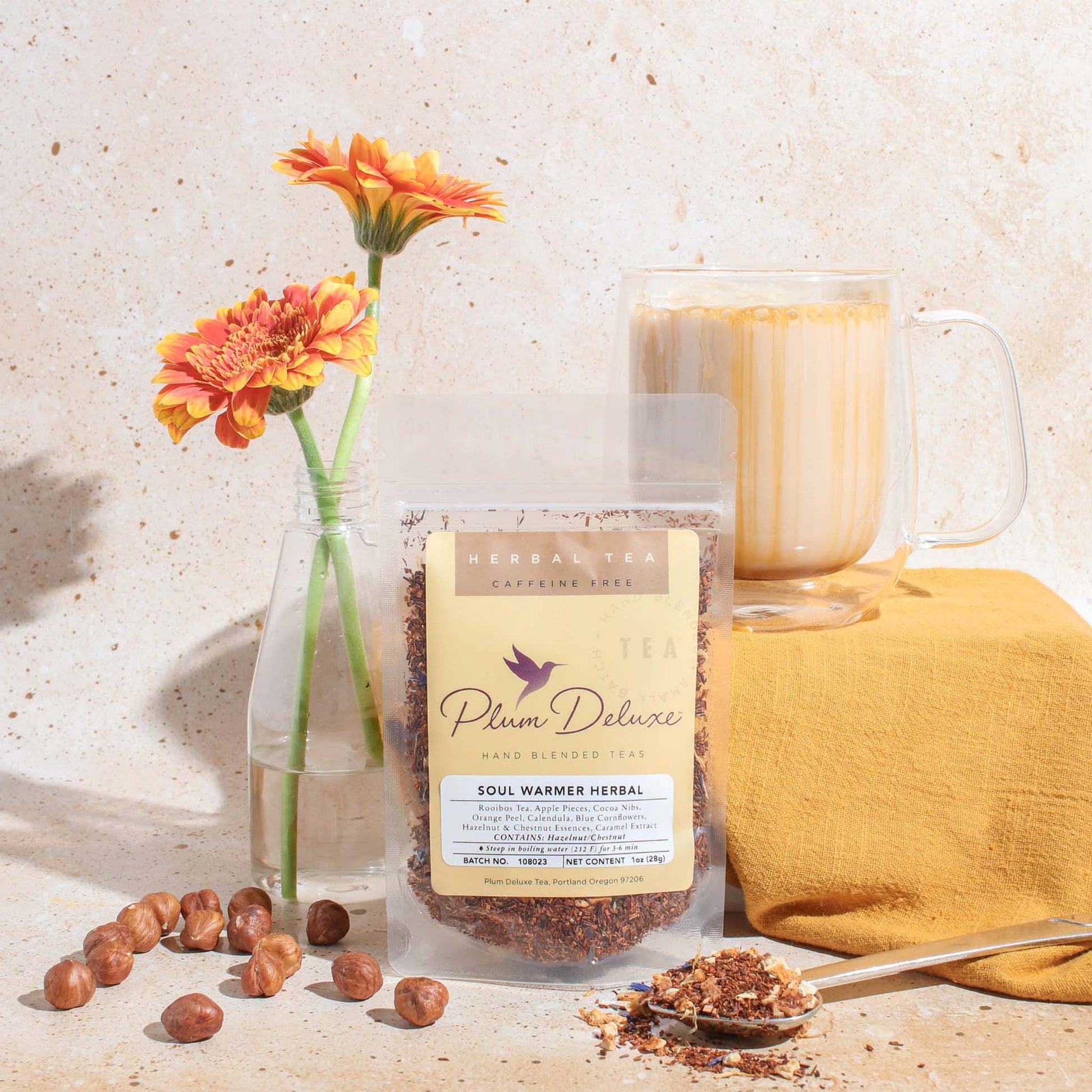 A 1oz package of Plum Deluxe "Soul Warmer" herbal tea displayed with a glass of brewed tea, scattered hazelnuts, and orange gerbera daisies on a neutral textured surface.