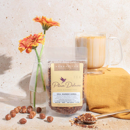 A 1oz package of Plum Deluxe "Soul Warmer" herbal tea displayed with a glass of brewed tea, scattered hazelnuts, and orange gerbera daisies on a neutral textured surface.