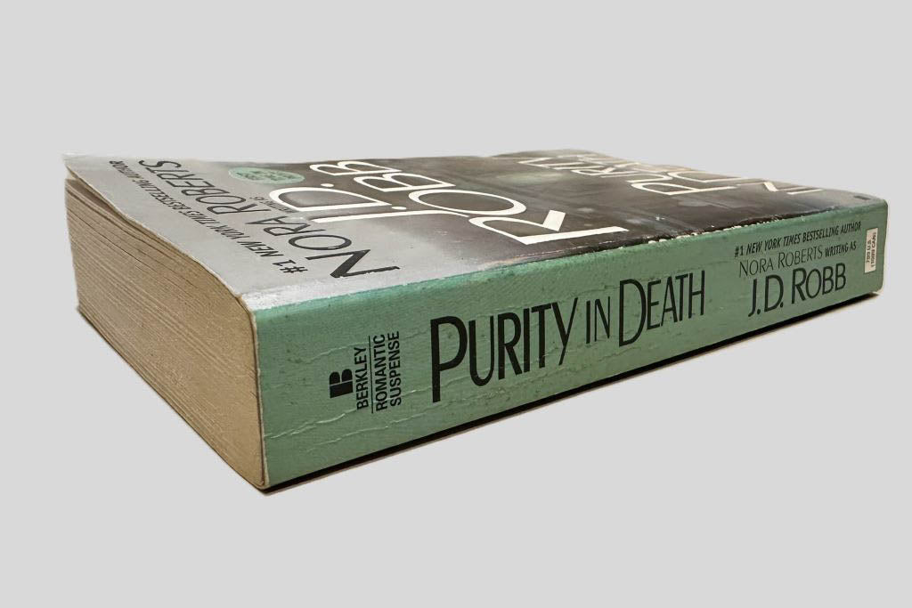 Close-up spine view of Purity in Death by J.D. Robb centered on a neutral light gray background. Spine shows minimal creasing. Pages lay flat.