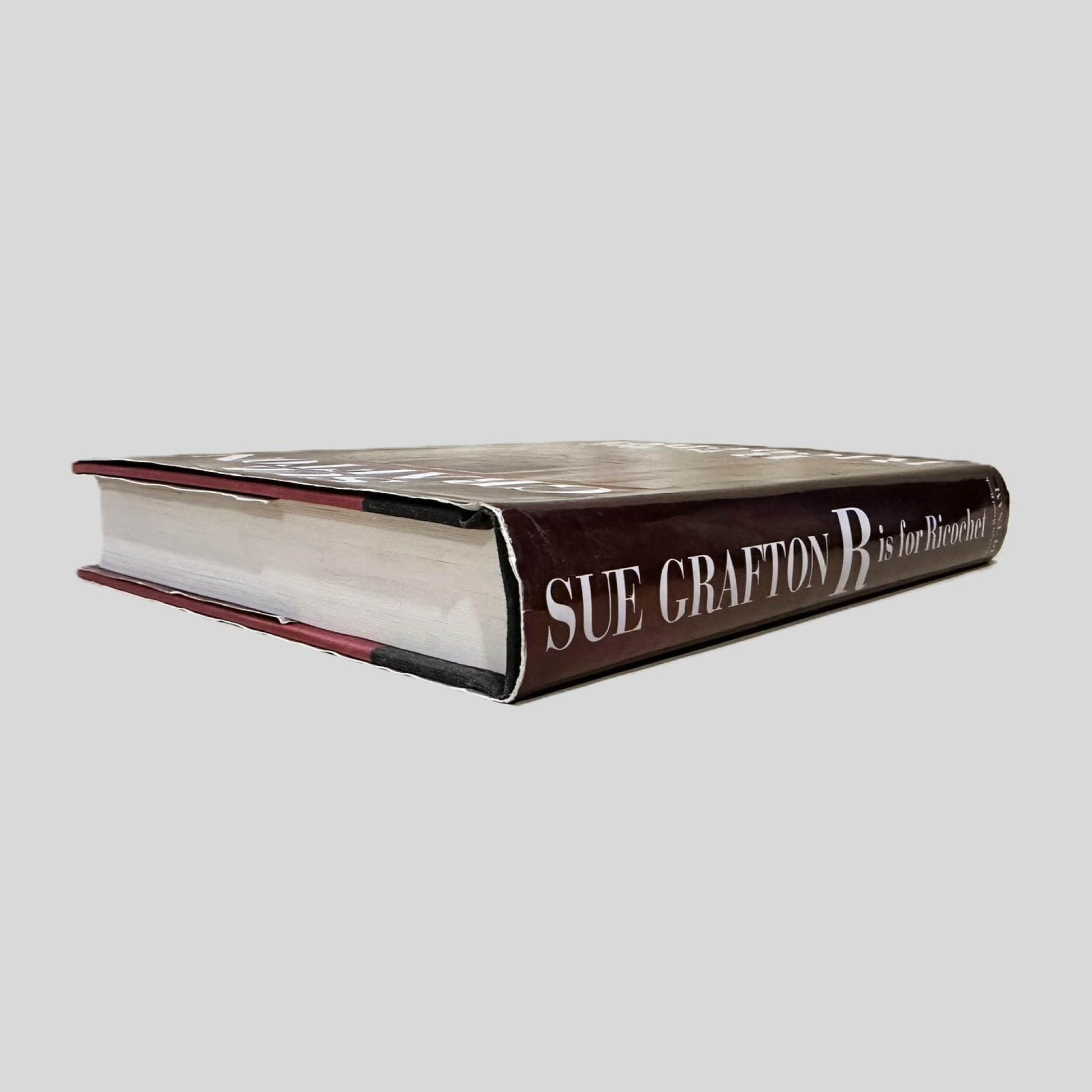 Close-up spine view of R is for Ricochet by Sue Grafton centered on a neutral light gray background. Dust jacket is present and undamaged.  Pages lay flat.