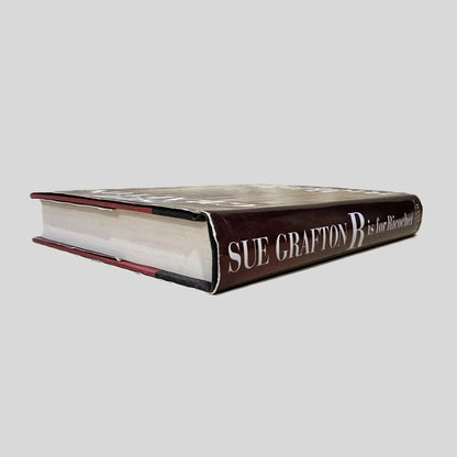 Close-up spine view of R is for Ricochet by Sue Grafton centered on a neutral light gray background. Dust jacket is present and undamaged.  Pages lay flat.