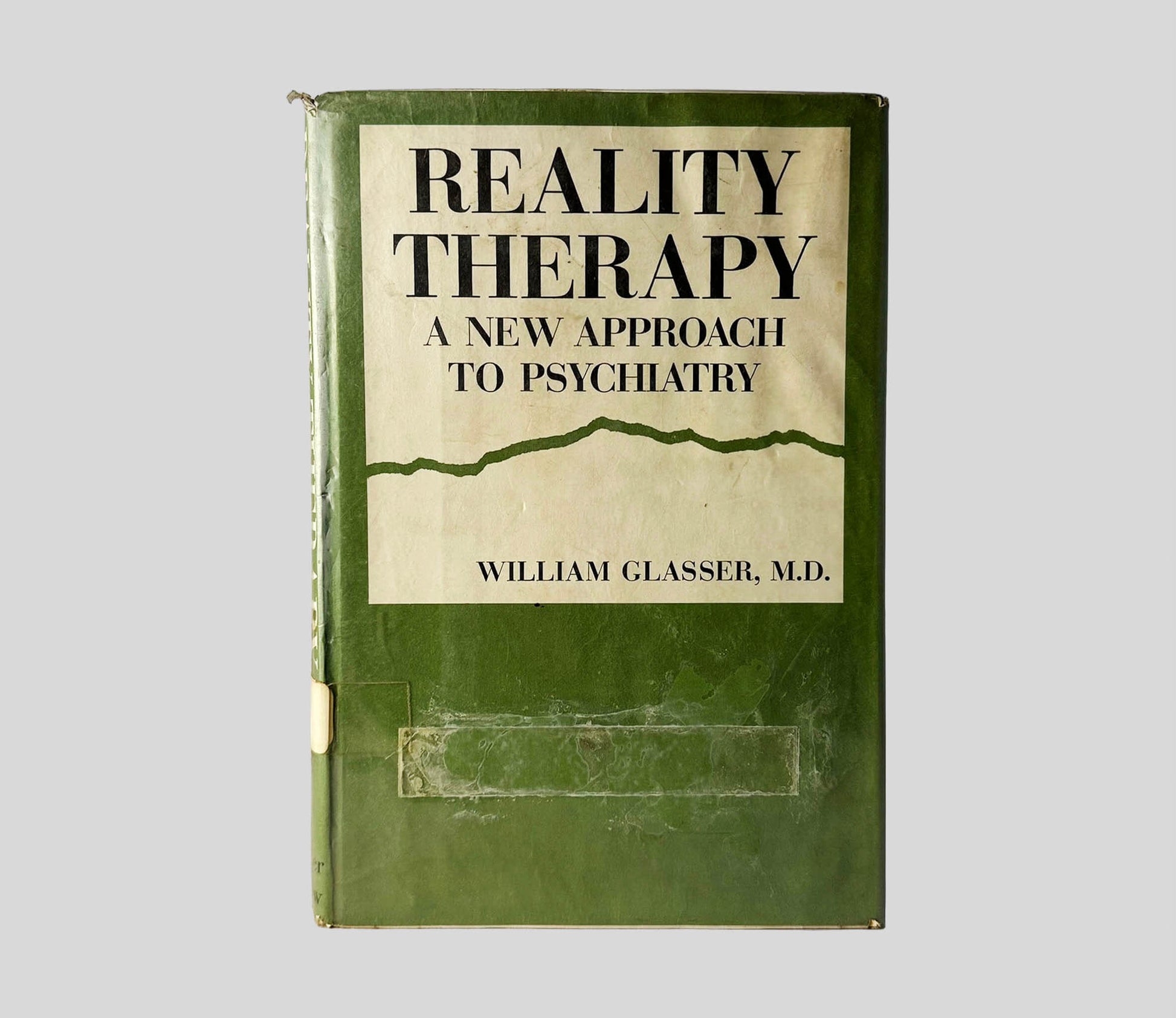 Front cover view of Reality Therapy by William Glasser, M.D. centered on a neutral light gray background. Dust jacket is present with library mylar cover. Transparent tape is attached on the lower end, below the title block. Top spine corner shows fraying.