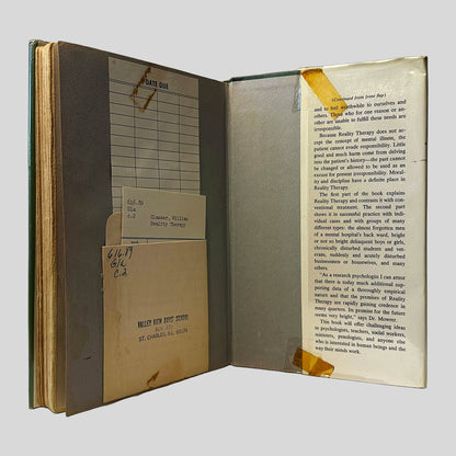 Inside back cover view of Reality Therapy by William Glasser, M.D. centered on a neutral light gray background. Library card pocket is attached to the back page from "Valley View Boys School" and the card is included. Dust jacket is present and is reinforced with tape. 