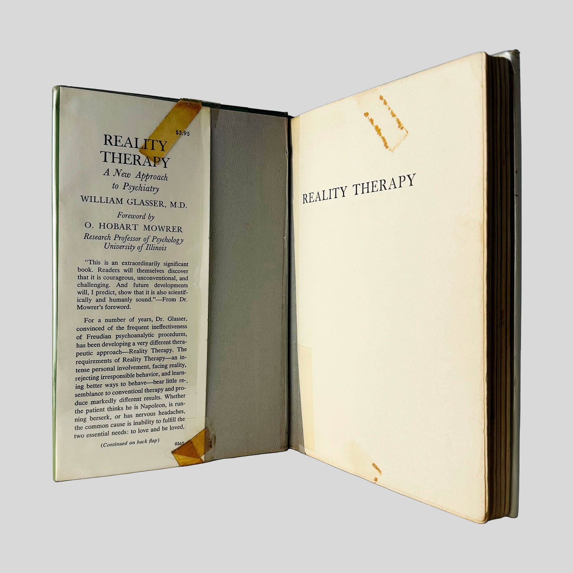 Inside front cover view of Reality Therapy by William Glasser, M.D. centered on a neutral light gray background. Dust jacket is reinforced to stay on the book with tape that has aged. 